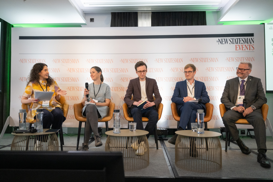 A backdrop featuring the new statesman logo and a panel of 5 individuals, on the left are two women in smart dresses, one is gesturing and speaking, the other is holding a microphone. The other panel members are three men all in suits, brown, blue, grey and glasses.