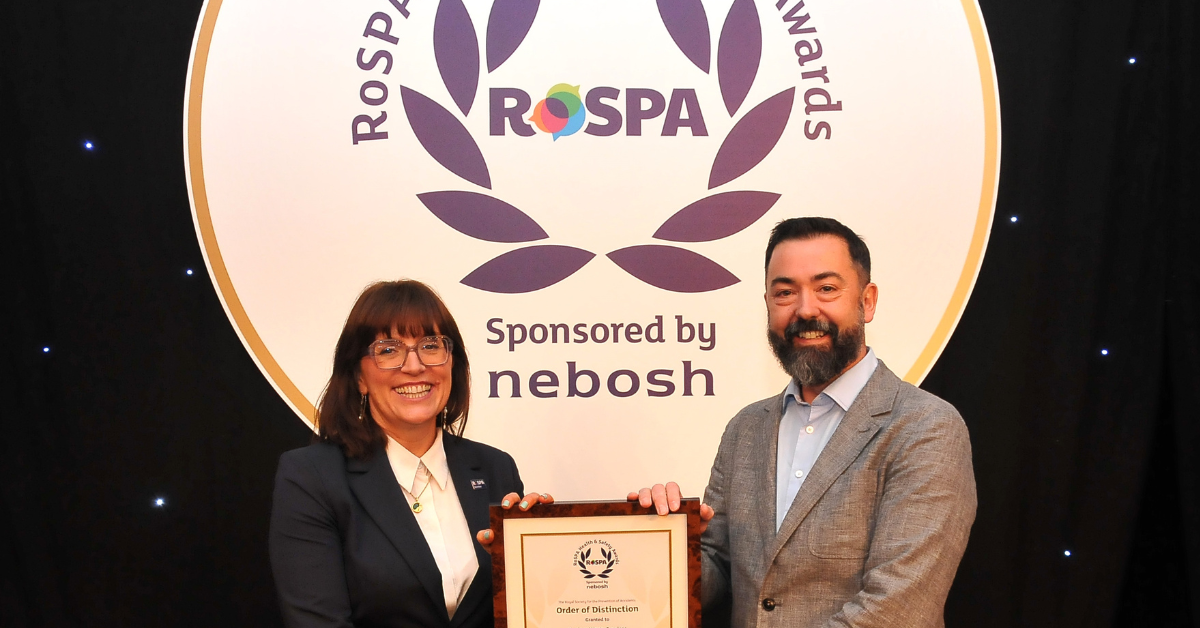 A smiling man receiving a framed award from a smiling woman in front of a branded backdrop
