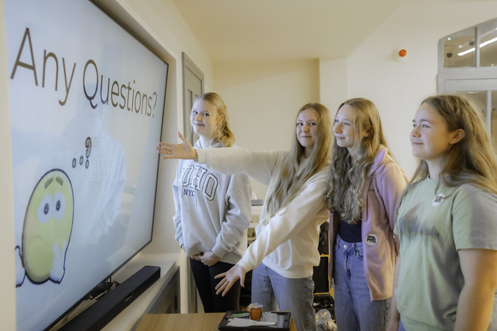 A group of 4 female students, one student is pointing to the large screen in front of them which has a presentation slide on it that says "Any questions?"