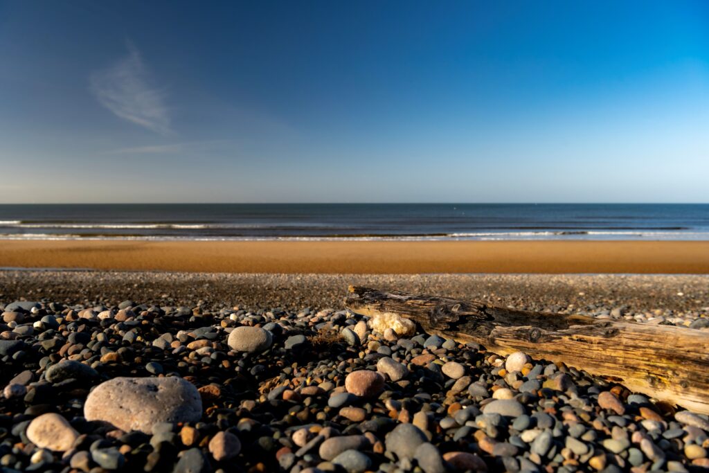 A view of the beach with the sea in the distance, a sandy beach and large pebbles and small log in the foreground.