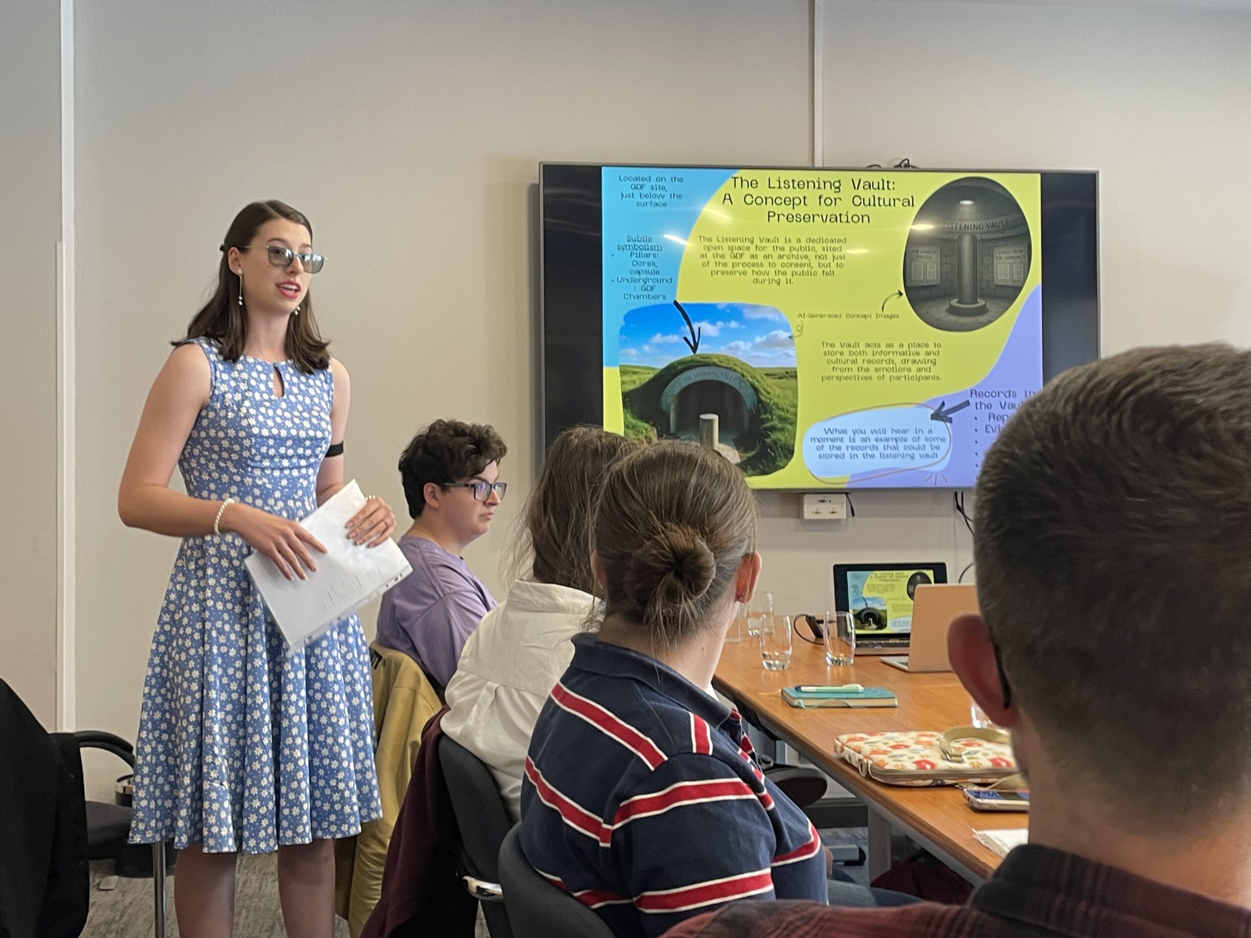 A young woman stood up in a light blue polka dot dress presenting a presentation on a large TV screen to a room of other young people who are sat at a meeting table.