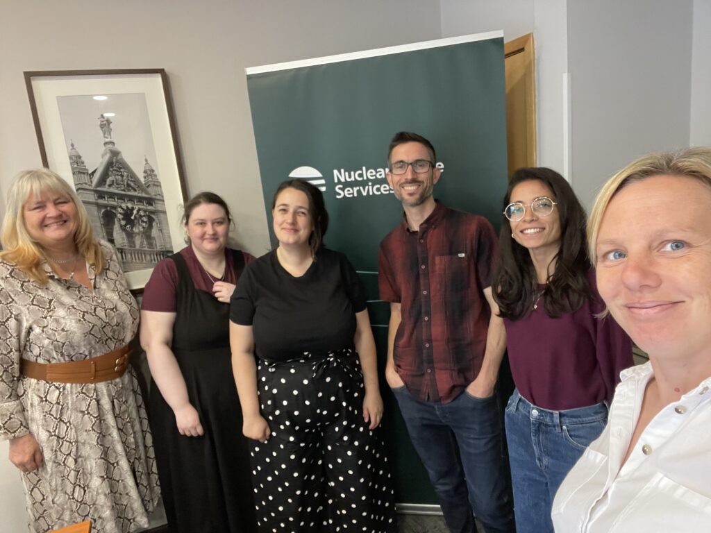 The delivery team of the national youth forum - a group of adults in smiling in front of a banner with the nuclear waste services logo