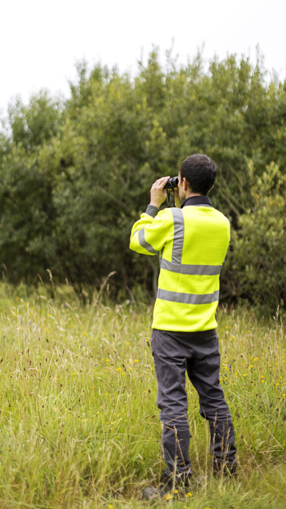 Man wearing a bright yellow high vis jacket, stood in a field and looking through binoculars.