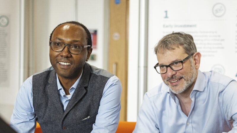 A man in a waistcoat with glasses, a man in a shirt with glasses and a beard are sat on a red sofa. They are smiling in between having a discussion.