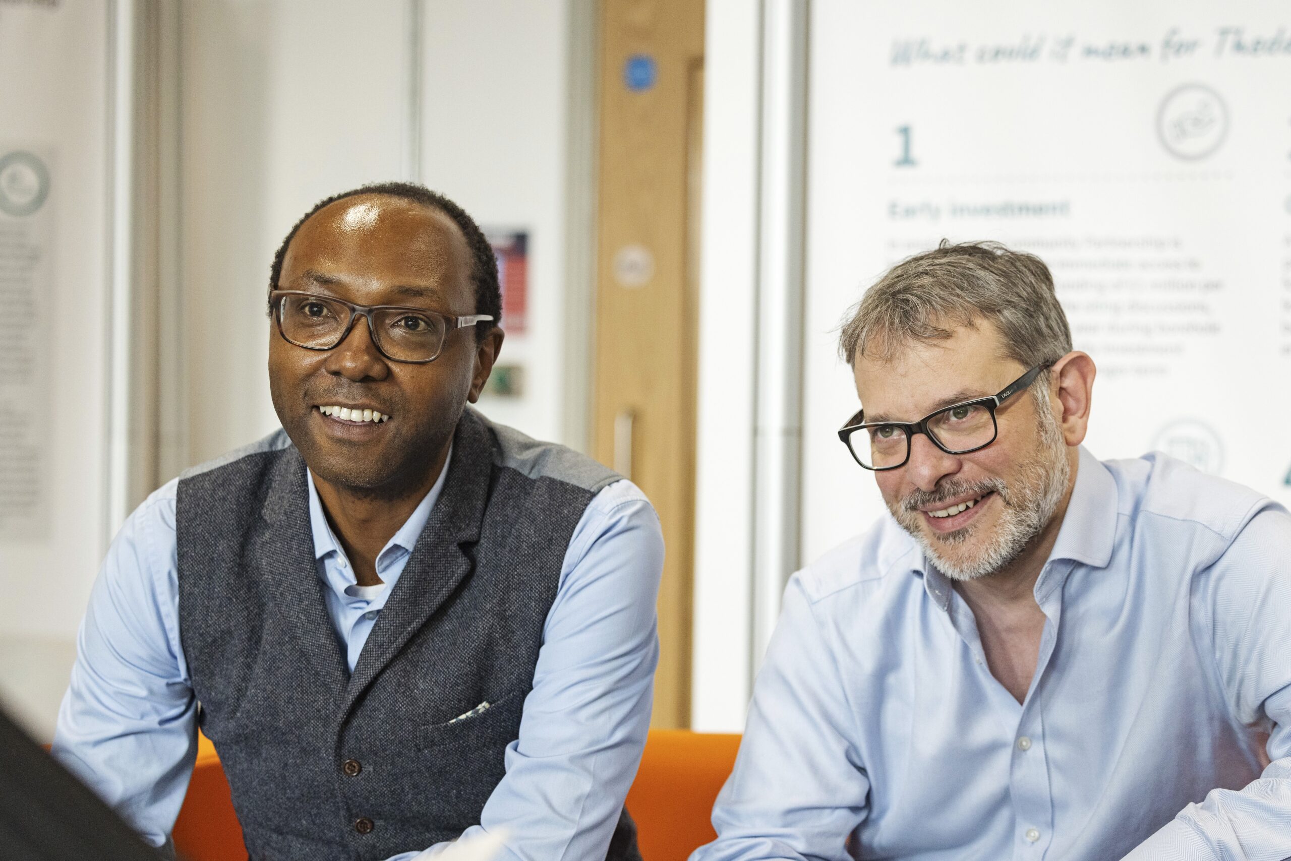 A man in a waistcoat with glasses, a man in a shirt with glasses and a beard are sat on a red sofa. They are smiling in between having a discussion.