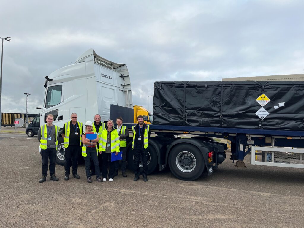 A group of six people, two women and 4 men all wearing hi vis yellow jackets stood together and behind them is a truck with a white cab and black covered load.