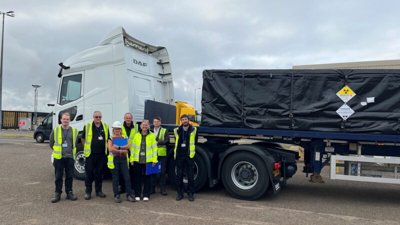 A group of six people, two women and 4 men all wearing hi vis yellow jackets stood together and behind them is a truck with a white cab and black covered load.