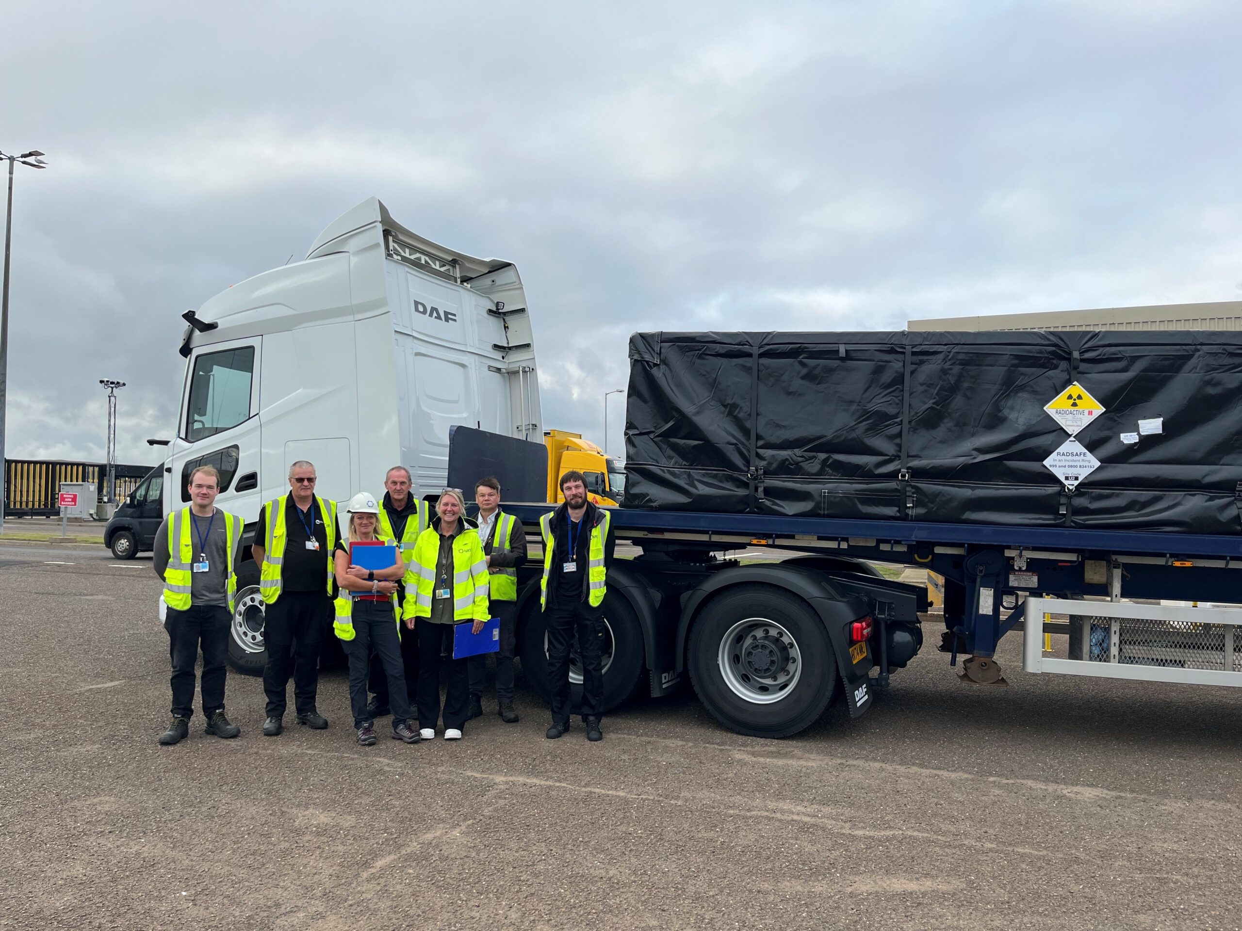 A group of six people, two women and 4 men all wearing hi vis yellow jackets stood together and behind them is a truck with a white cab and black covered load.