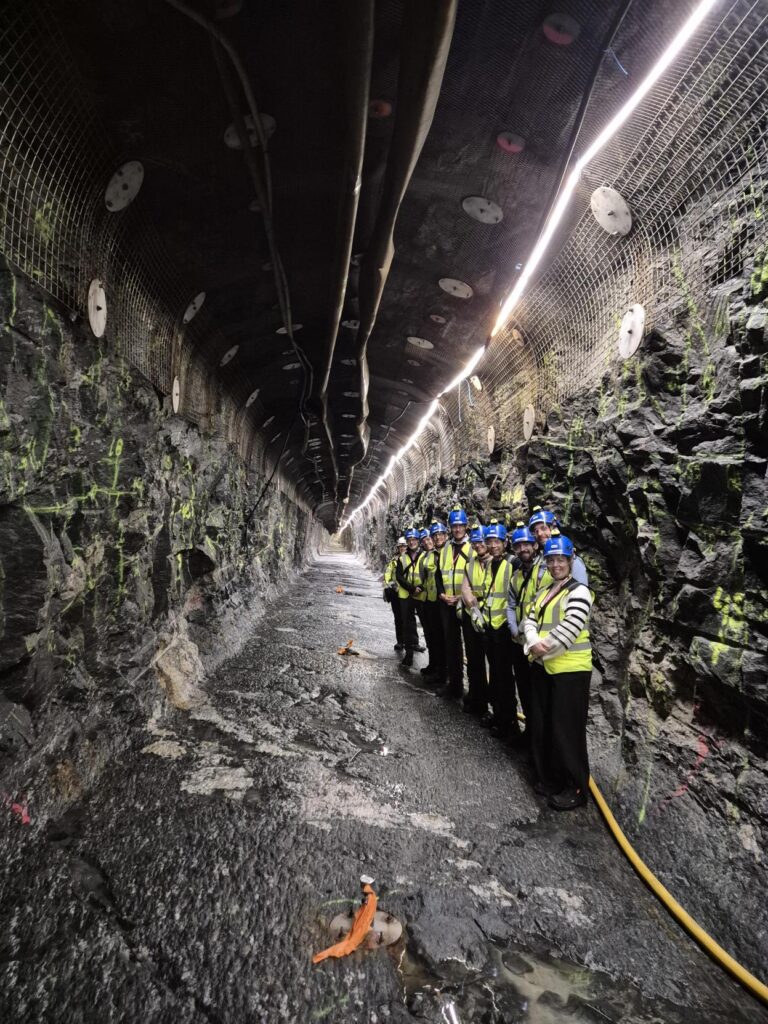 A group of 10 people wearing blue hard hats and yellow high vis jackets stood together in a line on the right hand side of a long underground tunnel in Onkalo, the Finnish geological disposal facility (GDF)
