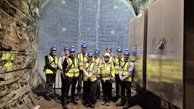 A group of 10 people wearing blue hard hats and yellow high vis jackets stood together in an underground facility in Onkalo, the Finnish geological disposal facility (GDF)