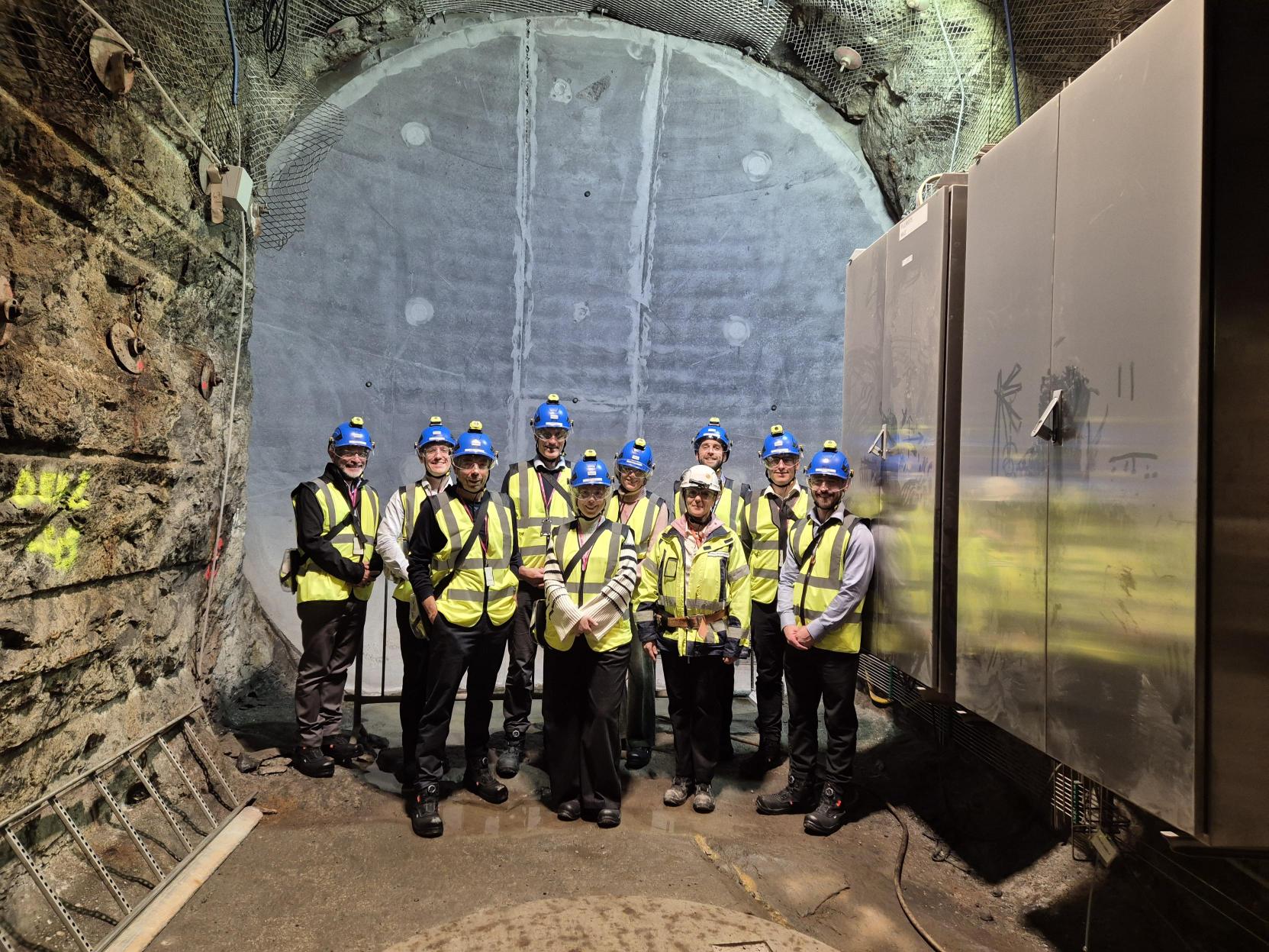 A group of 10 people wearing blue hard hats and yellow high vis jackets stood together in an underground facility in Onkalo, the Finnish geological disposal facility (GDF)