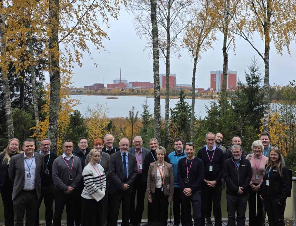 A group of 21 people stood in the foreground with a lake behind them and red buildings of Onkalo, the Finnish geological disposal facility (GDF) in the background.