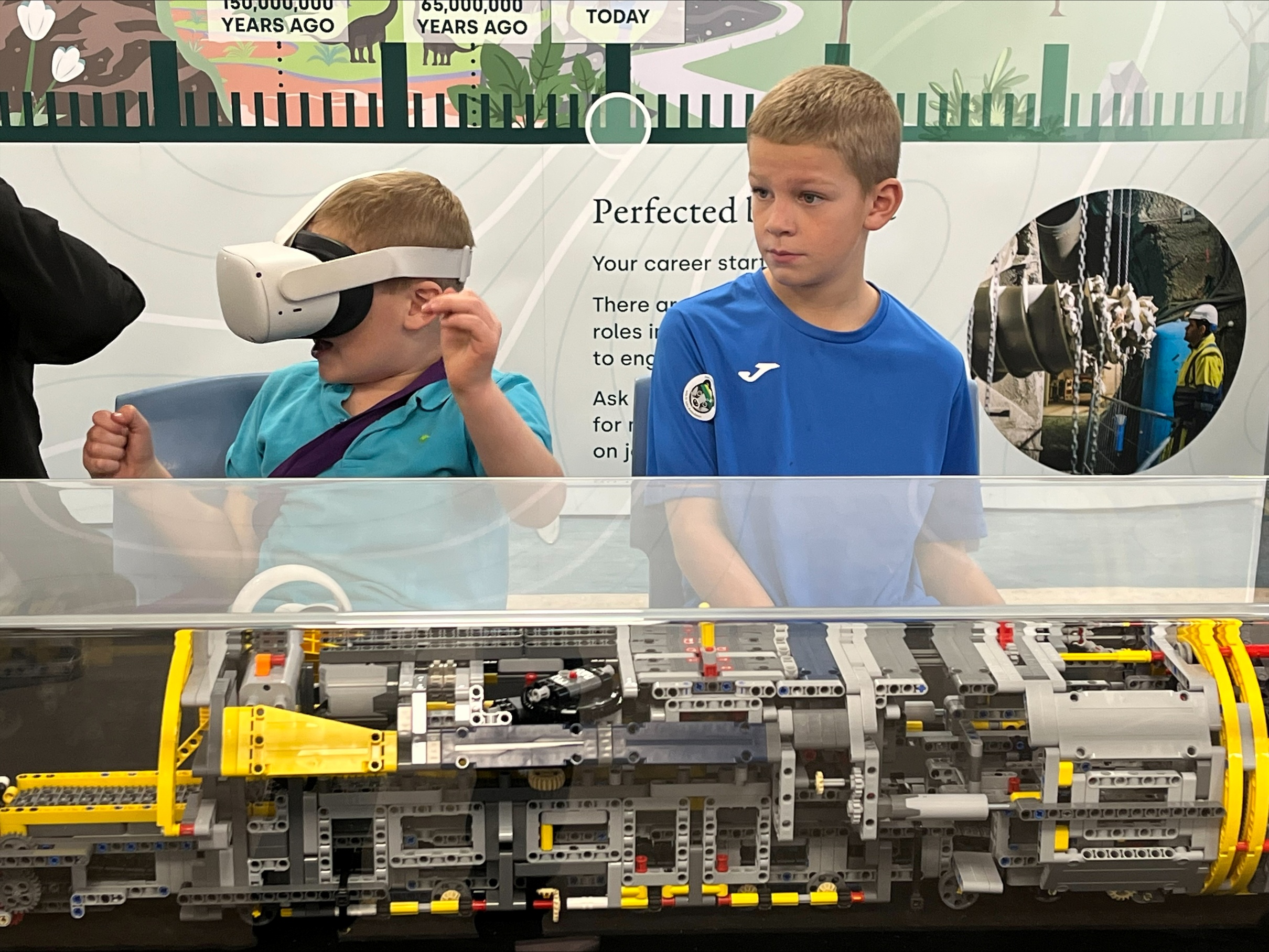 Two young boys stood next to a Lego model of a borehole drilling machine. One boy is wearing a VR headset.