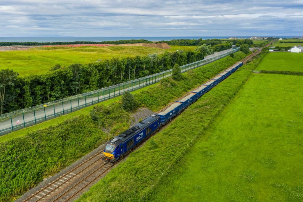 Train carrying aggregate on the track, green fields are either side and a fence on one side