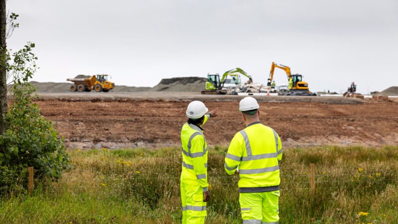 Two people wearing full Hi-Vis and hard hats, watching diggers carry out construction work.