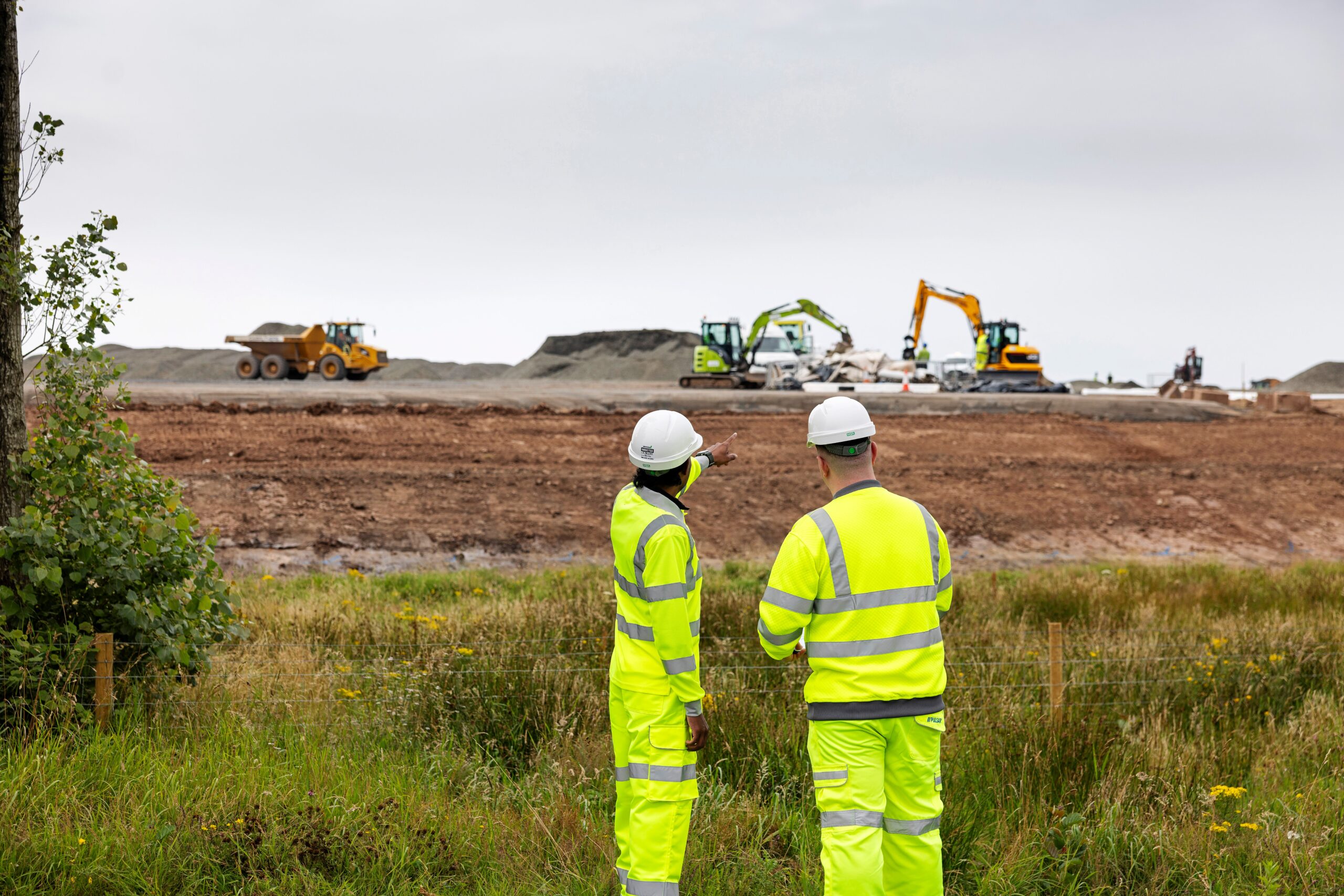 Two people wearing full Hi-Vis and hard hats, watching diggers carry out construction work.