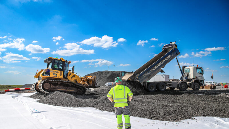 Man in high vis watching a lorry unload aggregrate in front of a digger