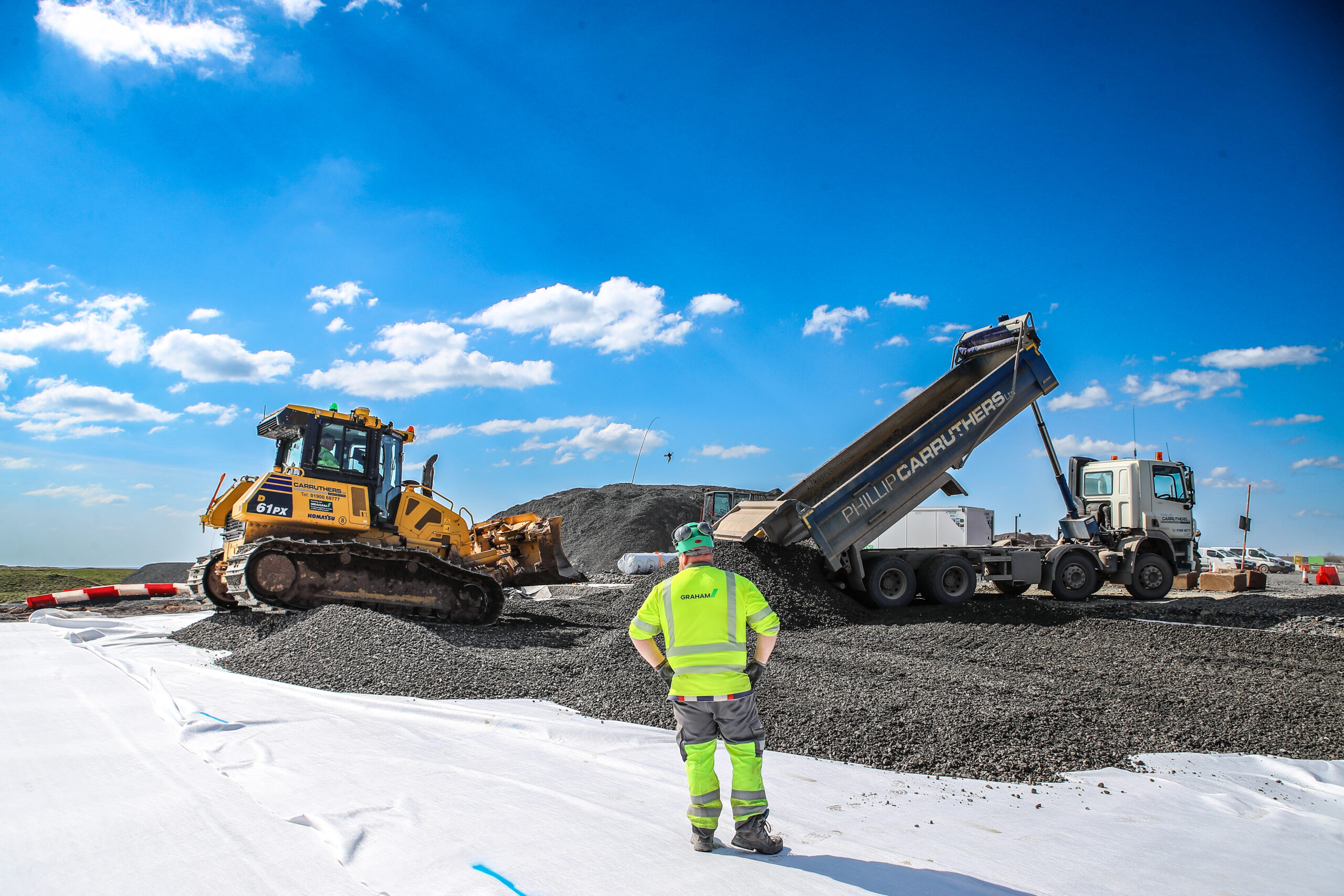 Man in high vis watching a lorry unload aggregrate in front of a digger