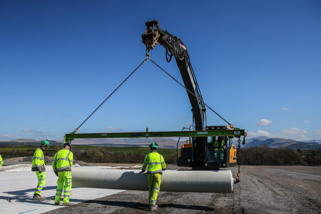 Three men in high vis clothing and hard hats walking in front of a small crane laying a large, white roll of liner in an open area covered with small, light grey gravel. In the background there are trees and some mountains
