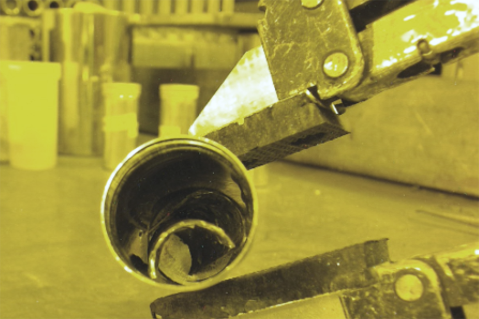 Fuel cladding handled remotely by a robotic arm inside a radioactive hot cell, viewed through protective lead-glass shielding